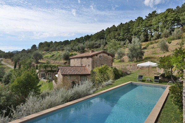 Une piscine bordée de pierres, un bâtiment en pierre avec un toit en tuiles et une zone herbeuse avec des arbres.