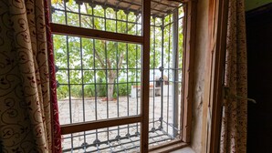 Una ventana con barrotes metálicos y marco de madera, cortinas con estampado floral, y vistas a árboles y un edificio.