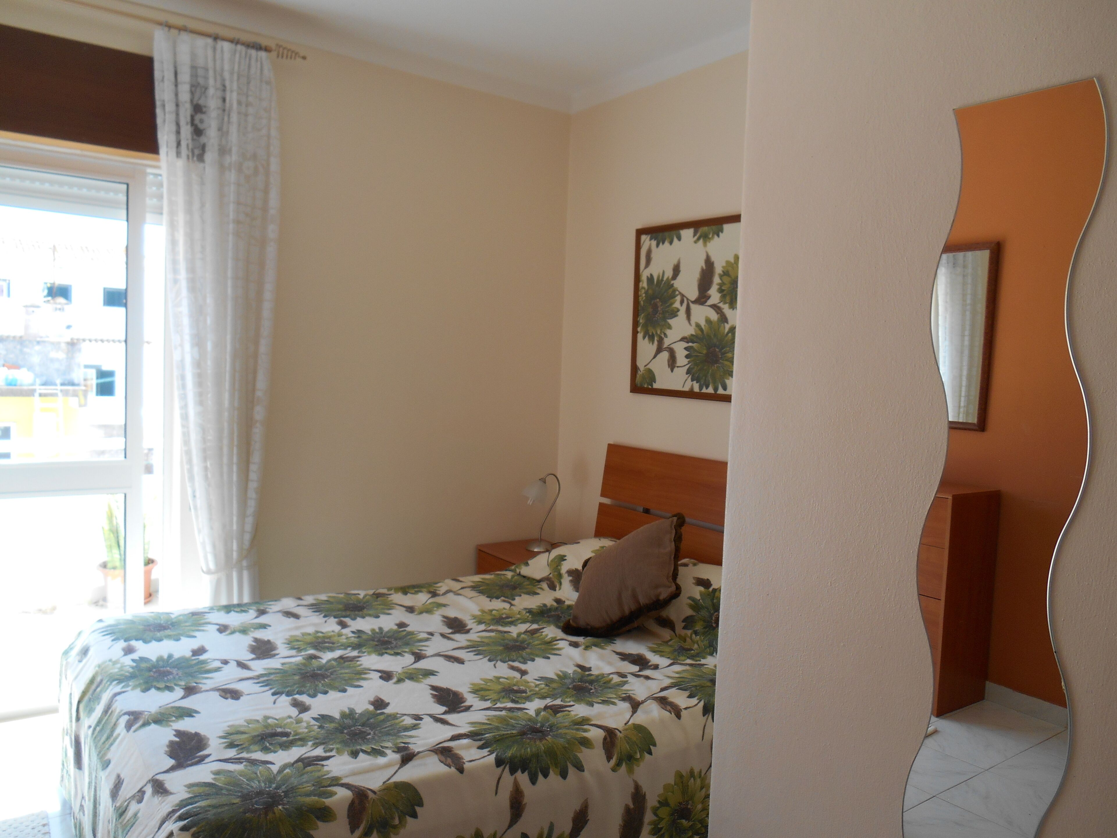 Une chambre à coucher comprenant un lit, une table de chevet, un miroir fixé au mur et une fenêtre avec des rideaux.