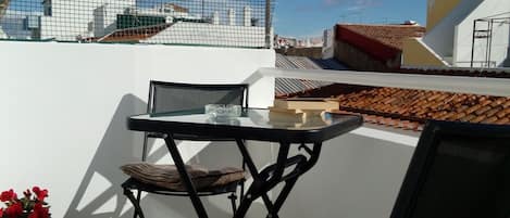 En terrass med ett bord och stolar, en krukväxt och utsikt över byggnader mot en klar himmel.