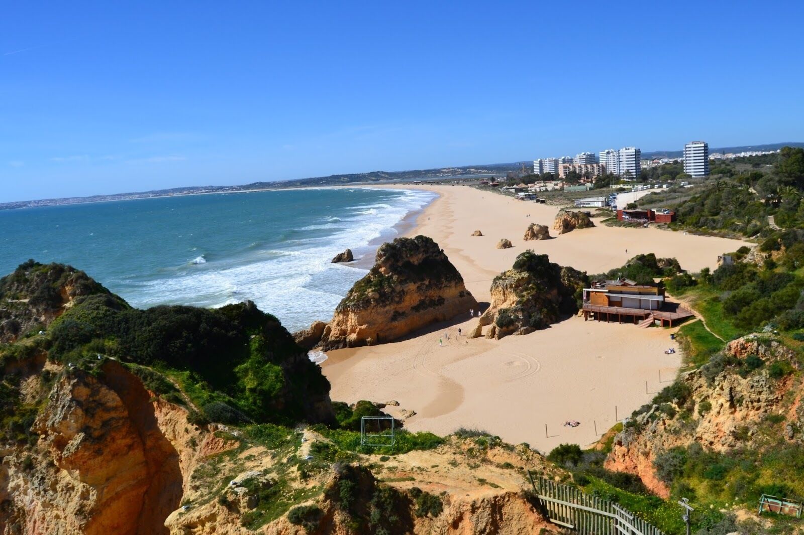 A coastal beach with rocky outcrops, a sandy beach, and a few buildings in the background.