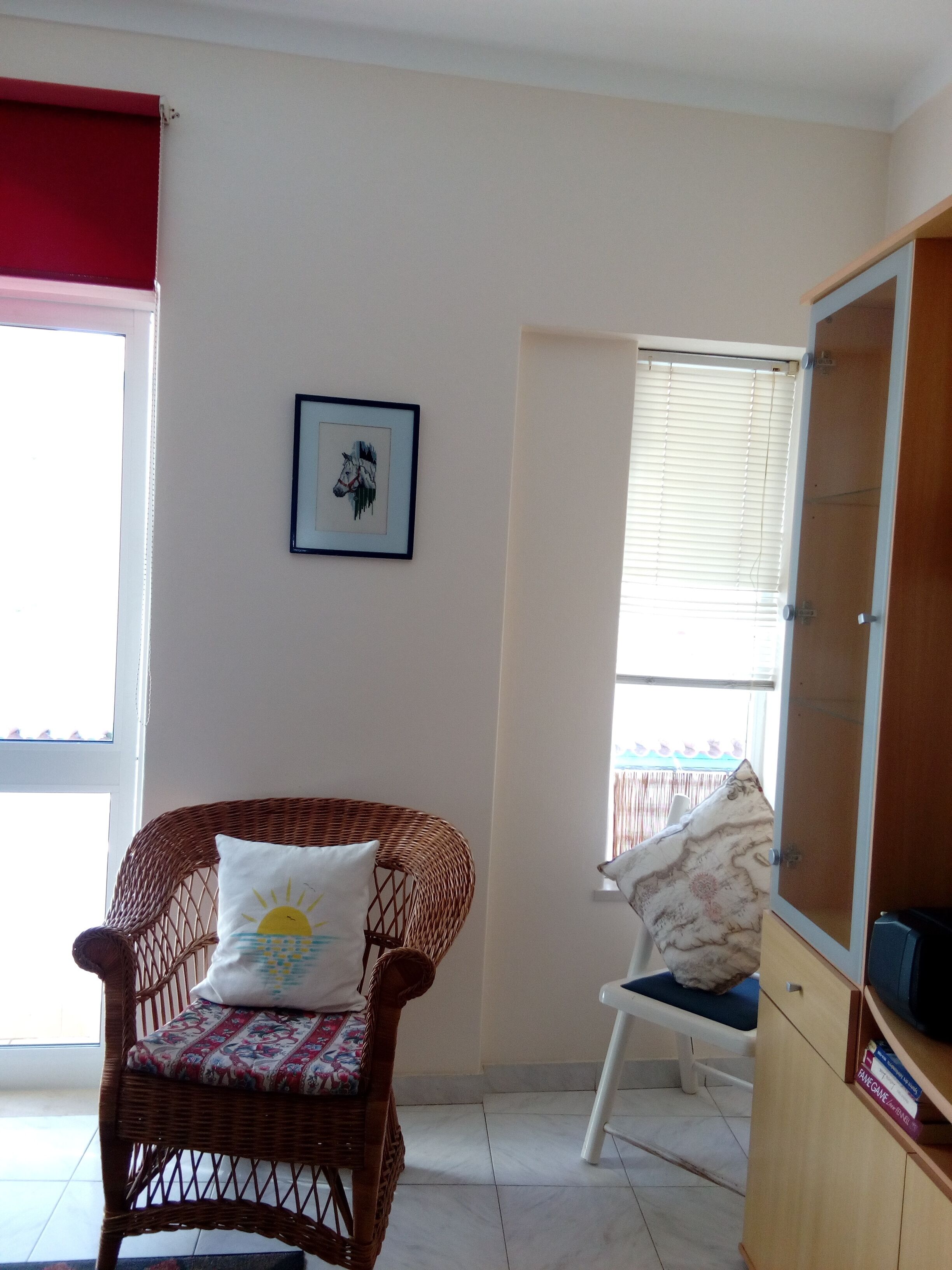 A room with a wicker chair, a white chair, a wooden cabinet, and a framed picture on the wall.