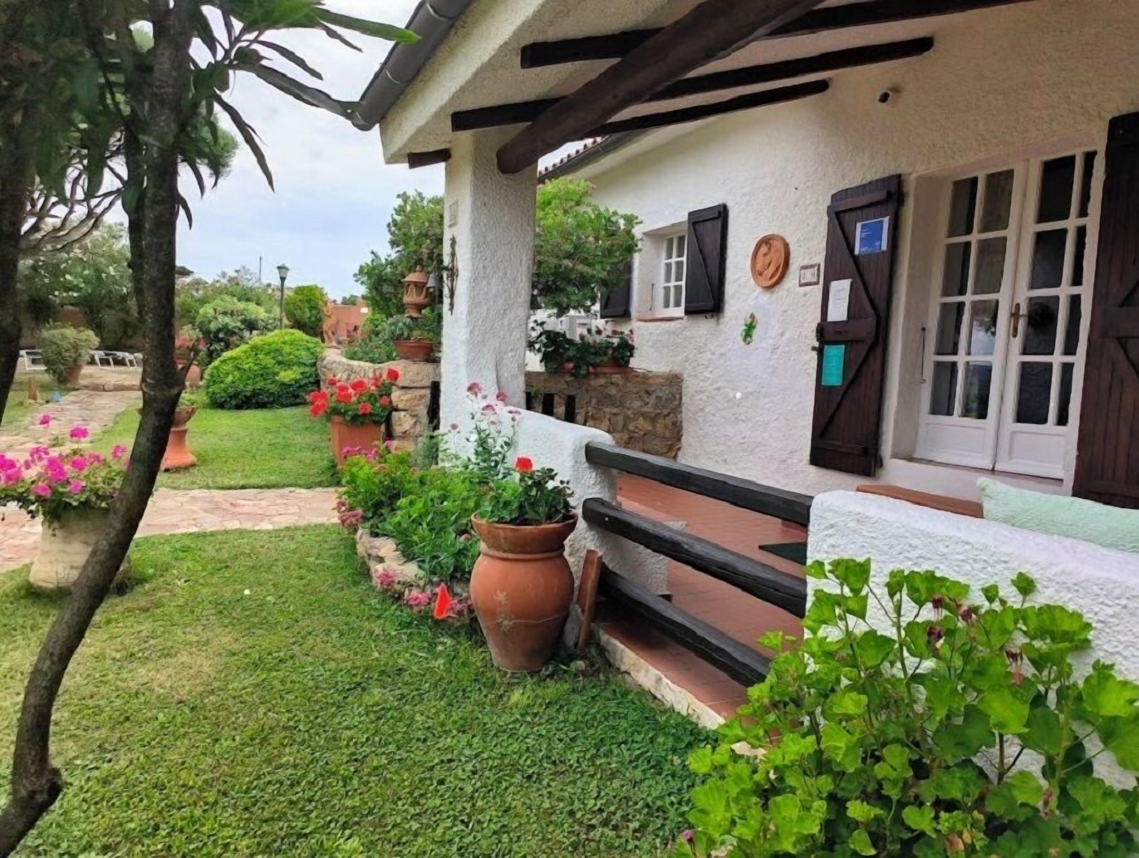 Una casa rústica con base de piedra, fachada blanca y porche de madera. Hay macetas con plantas y flores alrededor de la casa, y un camino de piedra que lleva a la entrada.