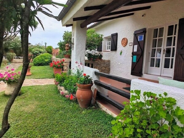 A rustic house with a stone foundation, a white exterior, and a wooden porch. There are potted plants and flowers around the house, and a stone pathway leading to the entrance.