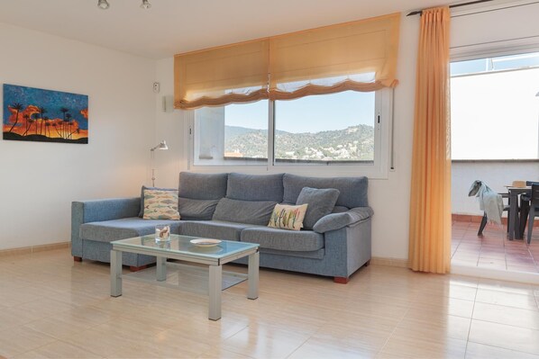 A living room with a sofa, a glass coffee table, a painting, and a view of the outside.