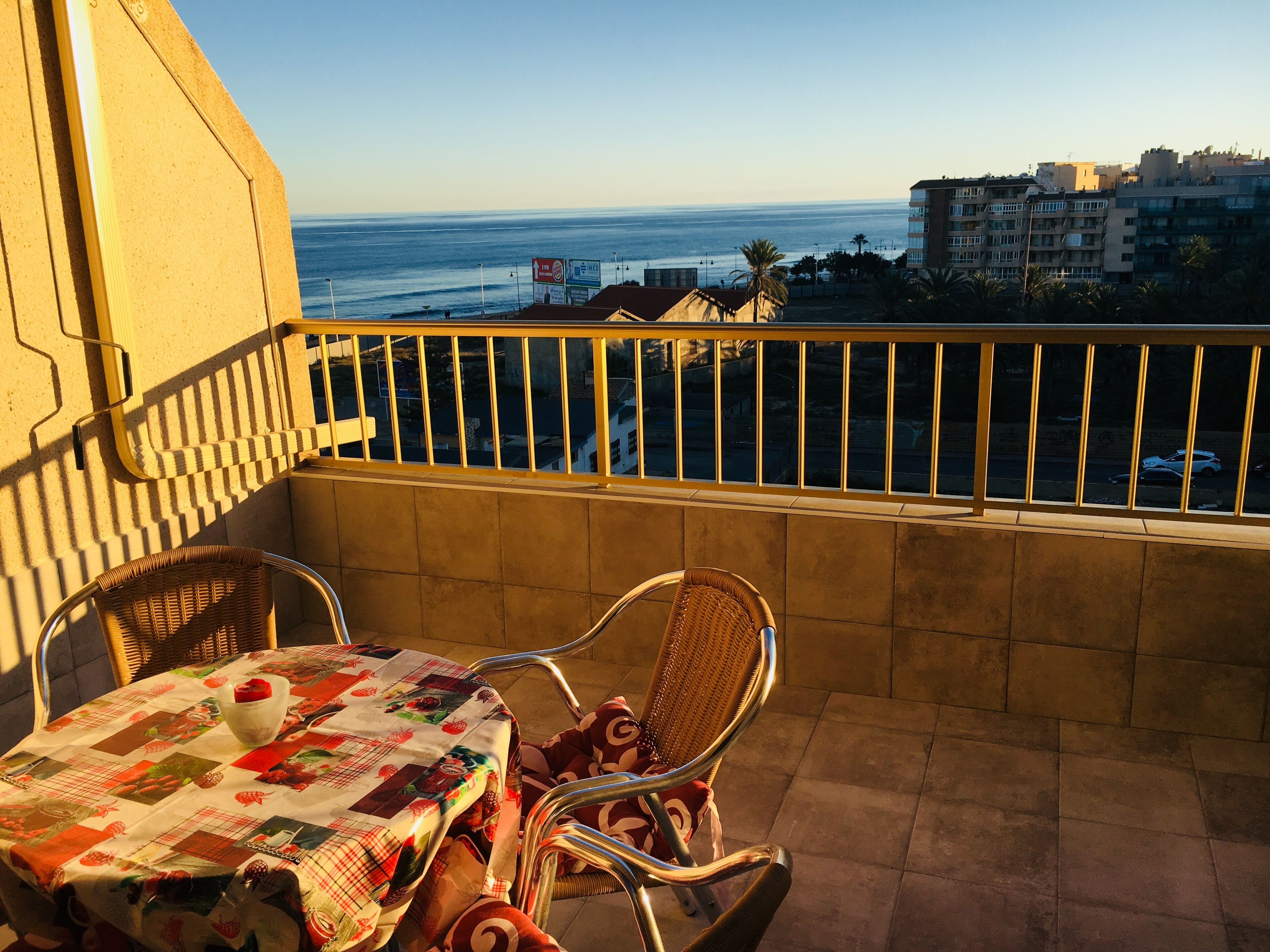 A balcony with a table and chairs, overlooking a cityscape and the sea.