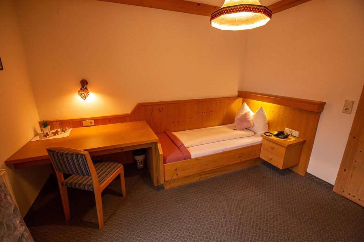 Une petite pièce agréable, dotée d’un bureau en bois, d’un lit simple, d’une chaise et d’une table de chevet avec une lampe.