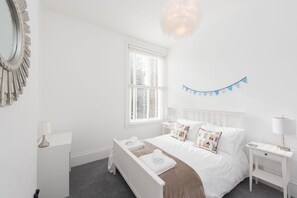 Une chambre à coucher blanche, avec un lit, des tables de chevet, une fenêtre avec des stores et une guirlande décorative de drapeaux.