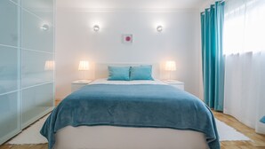 Ein modernes Schlafzimmer mit einem großen Bett, zwei Nachttischen, einem bei einem Bett stehenden Tischchen, einer Glastrennwand und blaustichigen Vorhängen.