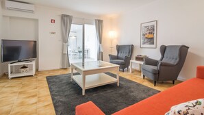 Ein modernes Wohnzimmer mit Flachbildfernseher, einem Glastisch, zwei grauen Sesseln, einem roten Sofa und einem gerahmten Bild an der Wand.