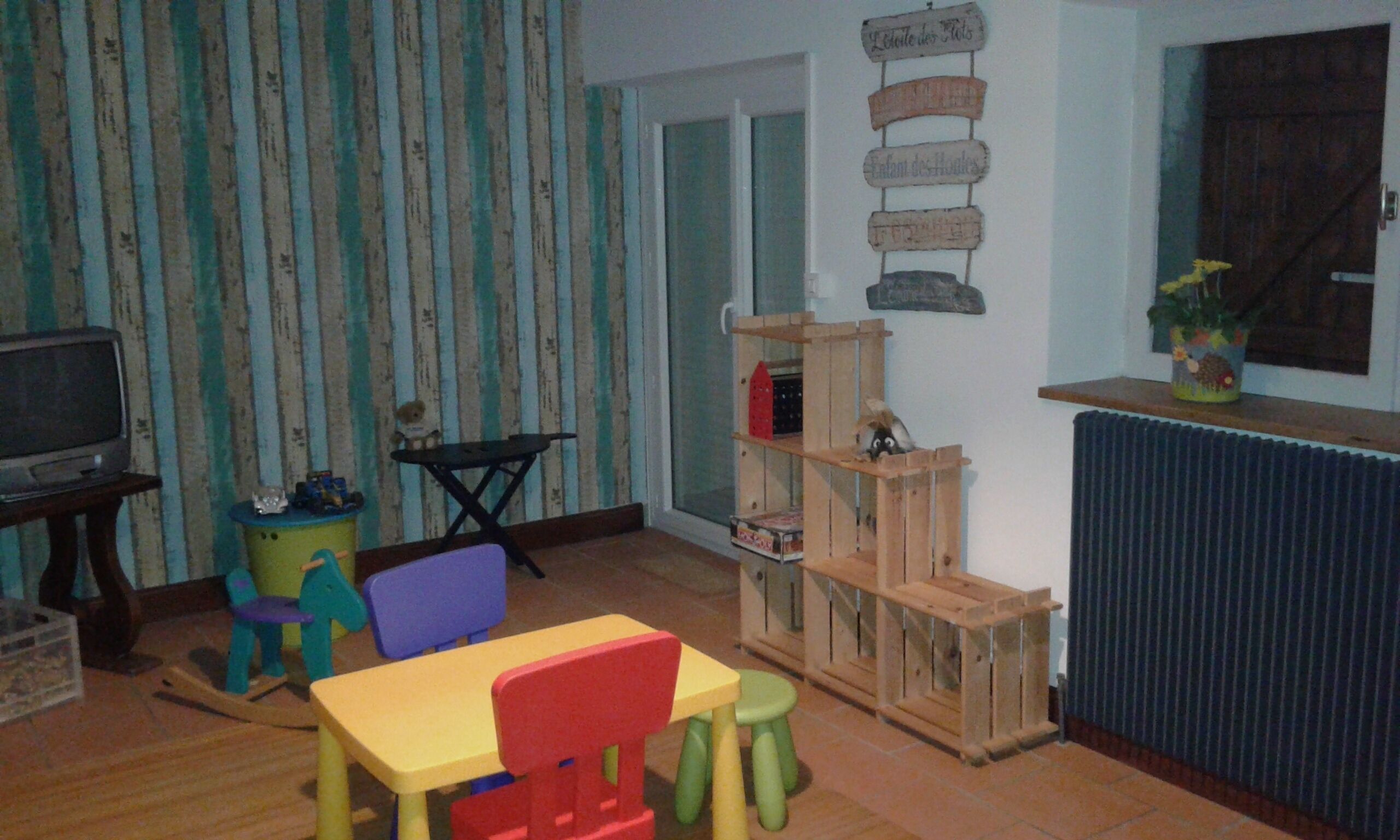 Une pièce avec un papier peint à rayures, une étag ère en bois avec des jouets, une télévision et une fenêtre.