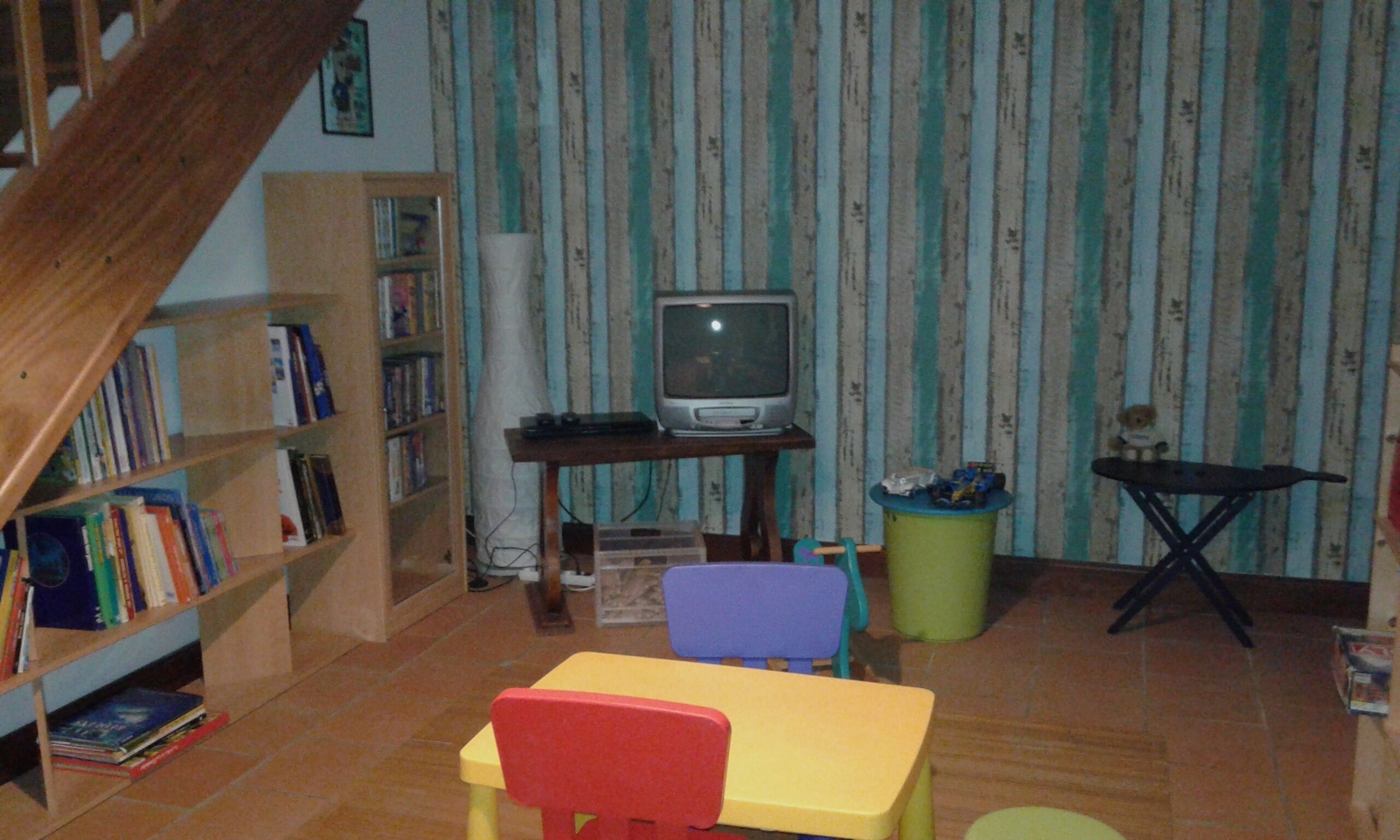 A room with a wooden staircase, a television on a stand, a bookshelf, a small table, and a trash bin.