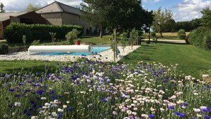 Piscina rodeada por um jardim com várias flores, uma casa com telhado inclinado e céu claro.