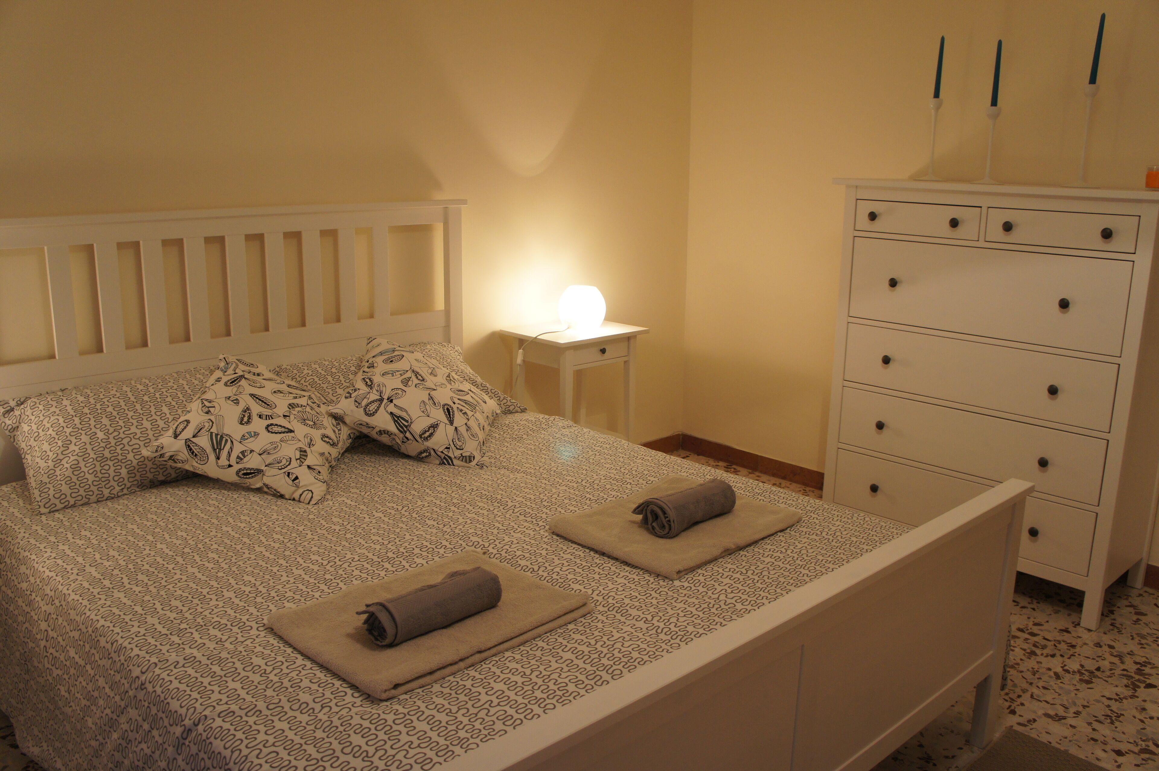 A neatly made bed with patterned bedding, a bedside table with a lamp, and a dresser with drawers.