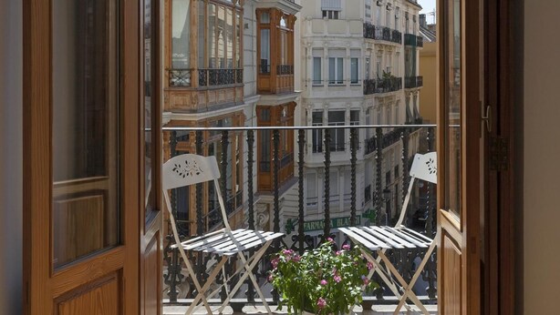 Ein Balkon mit zwei weißen Stühlen und einem Blumentopf, der auf eine Reihe historischer Gebäude mit aufwendigen Balkonen und großen Fenstern blickt.