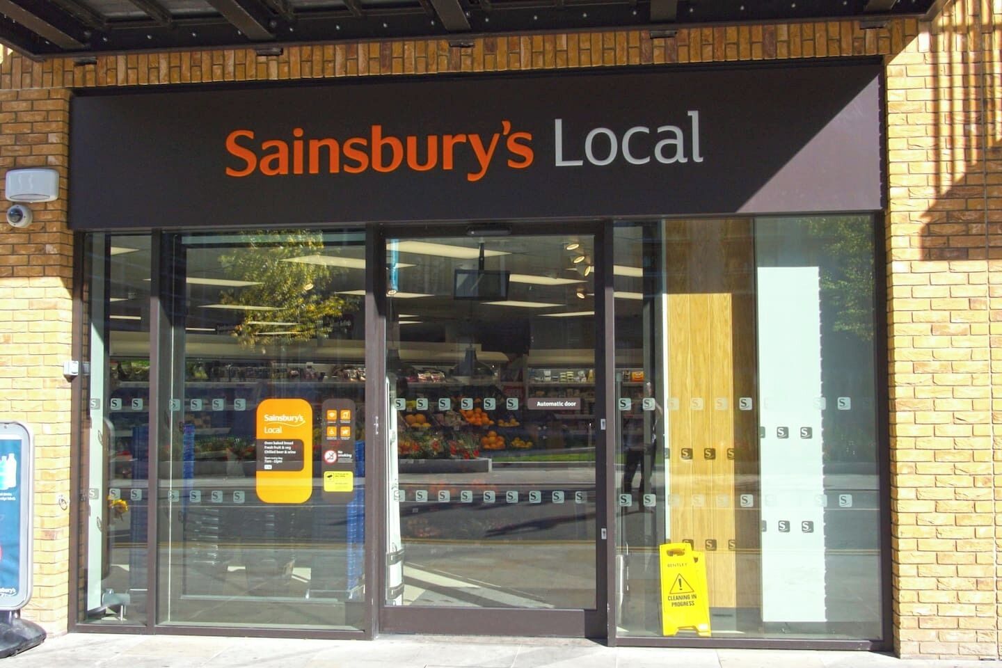 A Sainsbury's Local store with a clear glass front, automatic doors, and a sign indicating local produce.