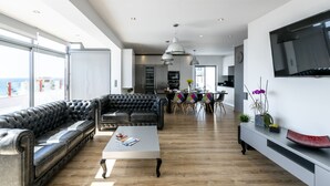 Un vaste espace de vie comprenant un canapé en cuir noir, une table basse blanche et un téléviseur fixé au mur.