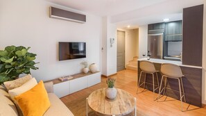 A modern living room with a sofa, coffee table, and a TV. There is a kitchen area with a dining set and bar stools. - Cozy apartment with terrace and free parking (Málaga)