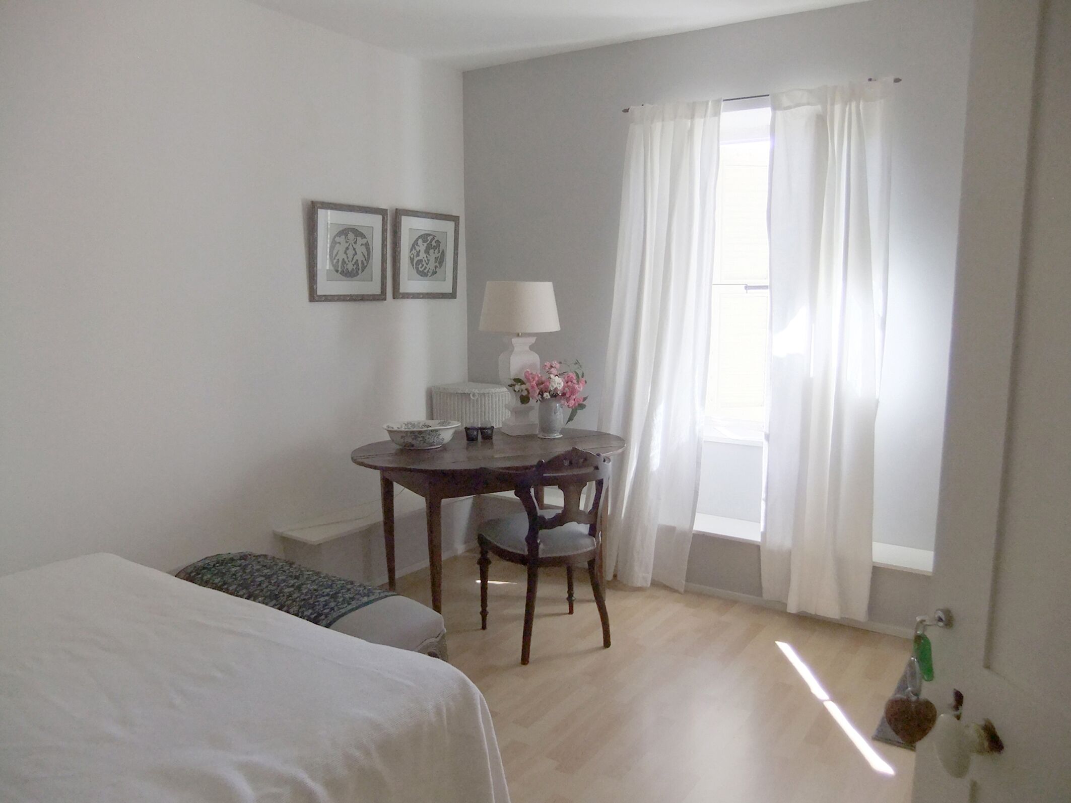 Habitación de hotel con una cama, una mesita de noche con una lámpara y flores, una silla y una ventana con cortinas.