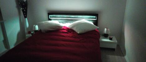 Un dormitorio con cama, almohadas, mesita de noche e iluminación en la pared.