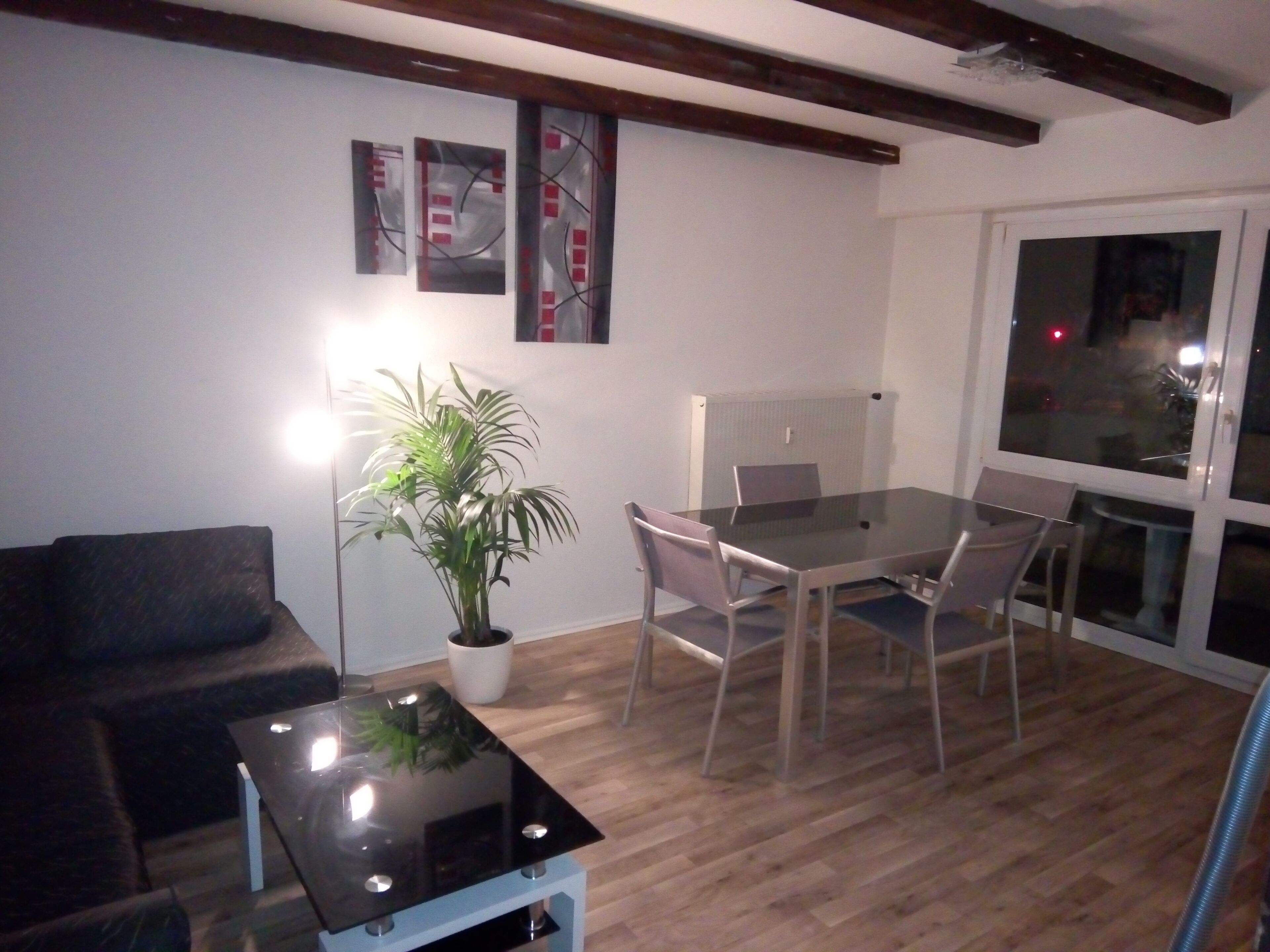 Ein modernes Wohnzimmer mit Glastisch, dunklem Sofa, Esstisch mit Stühlen, Topfpflanze und Bildern an der Wand.