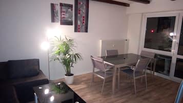 Ein modernes Wohnzimmer mit Glastisch, dunklem Sofa, Esstisch mit Stühlen, Topfpflanze und Bildern an der Wand.