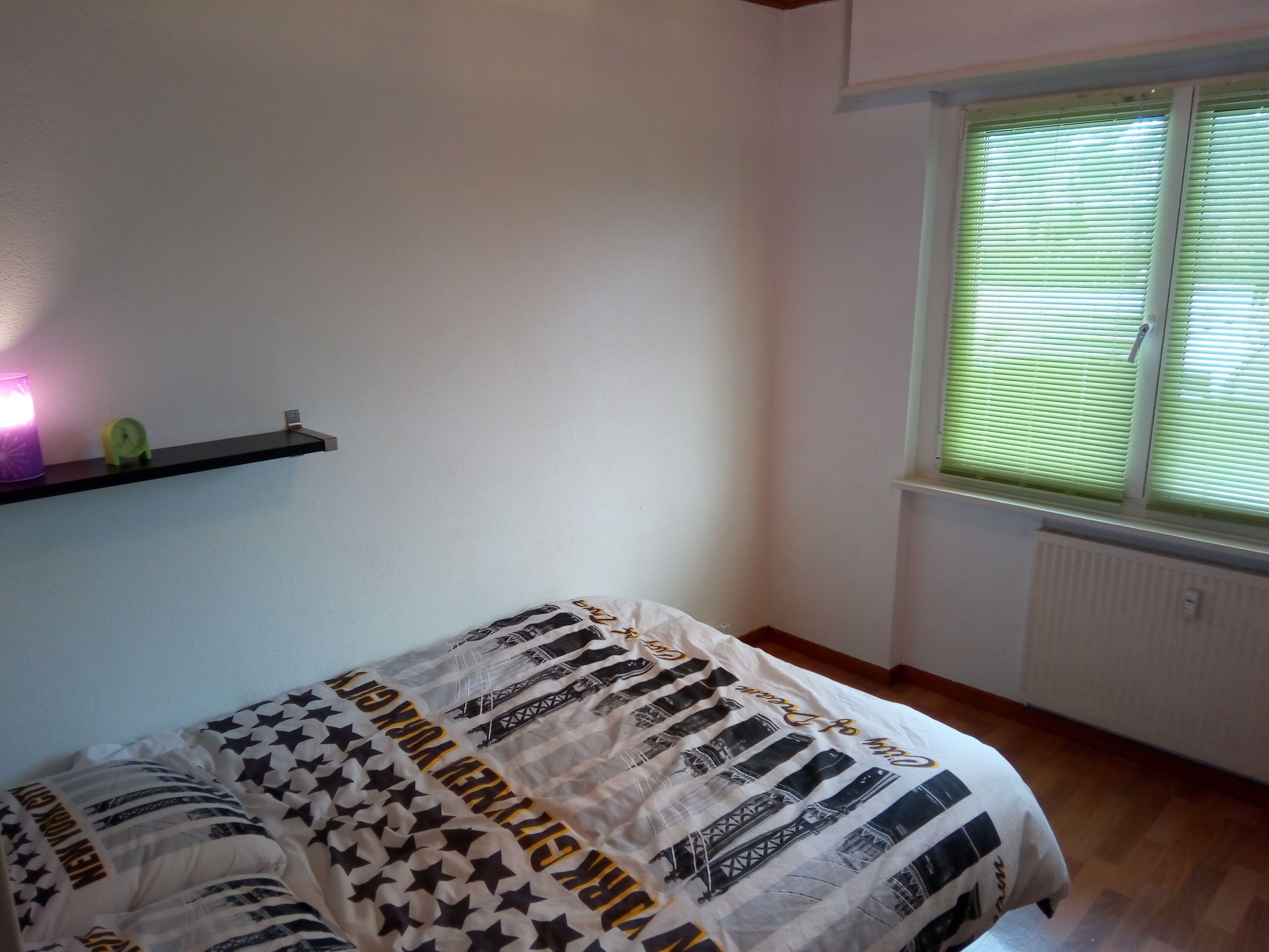 Quarto com cama, prateleira com abajur e janela com persianas.