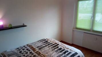 Quarto com cama, prateleira com abajur e janela com persianas.