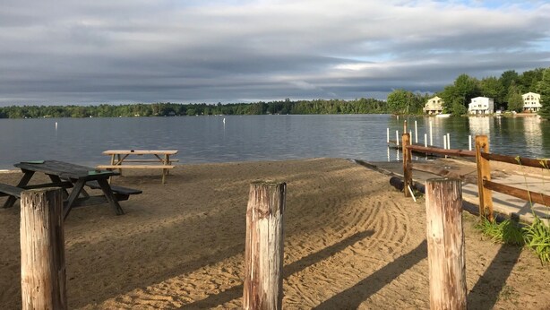 Blick auf einen See mit Sandstrand, Picknicktischen und einer Holzsteganlage.