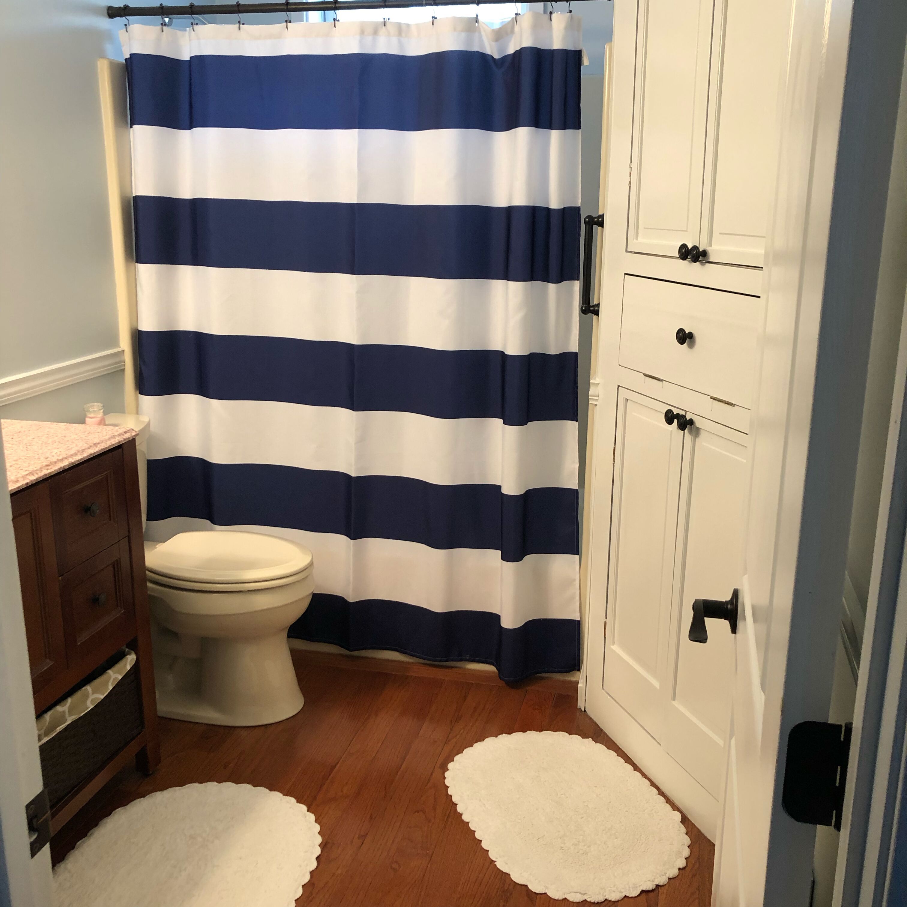 Ein Badezimmer mit gestreiftem Duschvorhang, Toilette, Holzschränken und zwei weißen Teppichen auf dem Boden.
