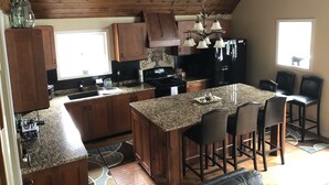 Eine Küche mit Holzausstattung, Granit Arbeitsplatten, einer zentralen Kochinsel mit Barhocker sowie einem schwarzen Kühlschrank und einer Pendelleuchte.