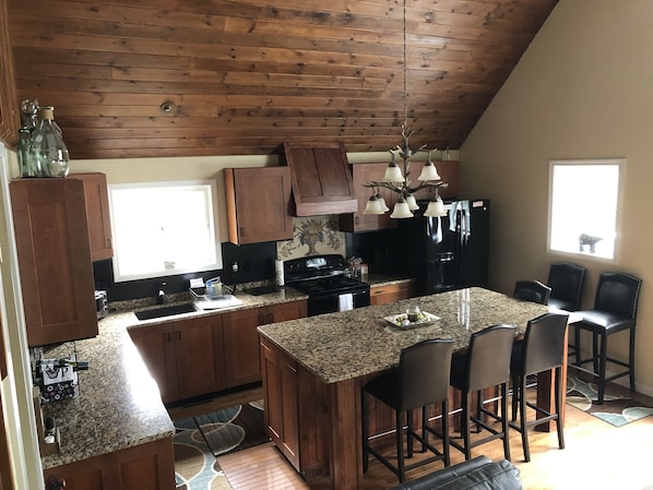 A kitchen with wooden cabinets, granite countertops, a central island with bar stools, a black refrigerator, and a hanging light fixture.