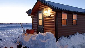 Eine Holzhütte mit Metall-Dach, umgeben von Schnee, mit Schneeschaufel und Eimer in der Nähe.
