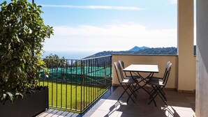 A balcony with a table and chairs, a potted plant, and a view of a green field and mountains. - Le Capannelle - Figaro by Feeling Italy (Massa Lubrense)