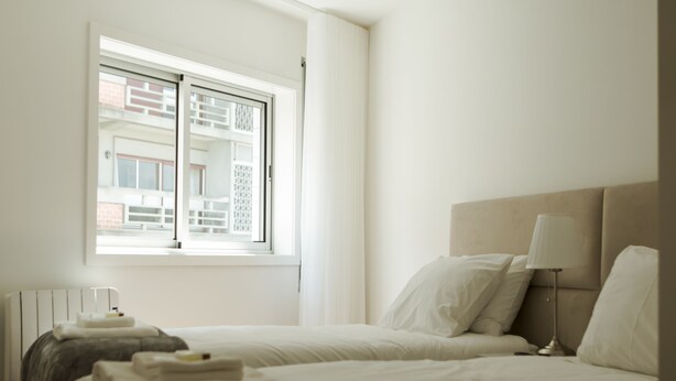A hotel room with two beds, a radiator, a window with a view of buildings, and a bedside table with a lamp. - Nightingale Apartments (Porto)