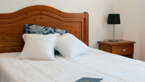 Un lit bien fait, avec des draps blancs et deux oreillers, dont l’un est orné d’un motif floral. Un livre est posé sur le lit.