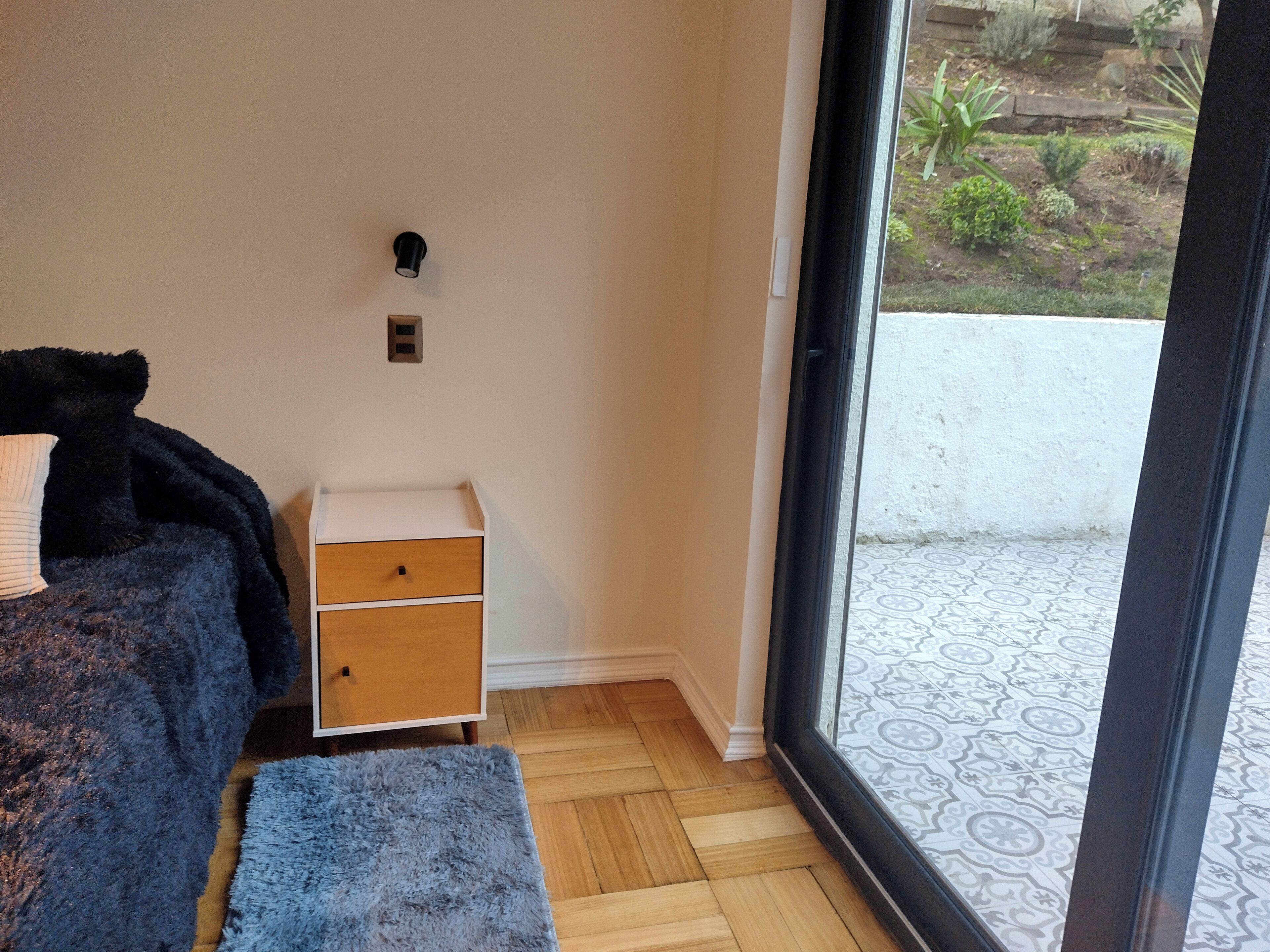 Ein Schlafzimmer mit einem Bett, einem Nachttisch, einer Schublade, einem flauschigen Teppich und Blick auf einen Garten durch eine Glastür.