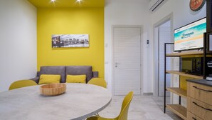 Un soggiorno moderno con un tavolo da pranzo rotondo, sedie gialle, un divano grigio con cuscini gialli, un mobile contenitore in legno e una televisione che mostra un sito web.
