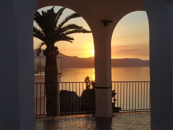 A balcony with a view of the sunset over a body of water, framed by architectural arches.