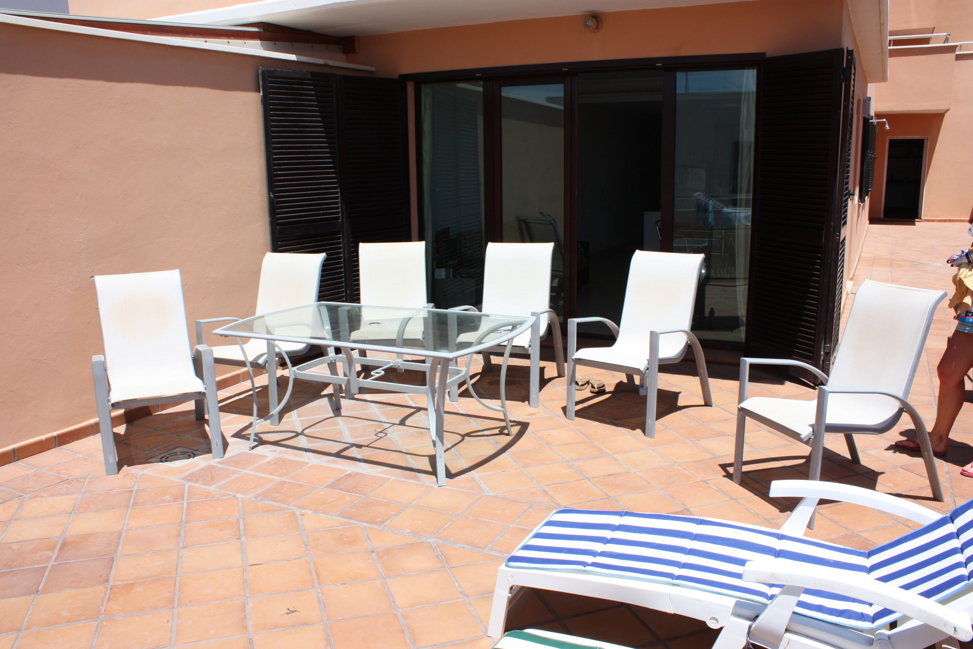 Um pátio externo com mesa e cadeiras de vidro, uma poltrona de lounge listrada e uma porta de correr de vidro.