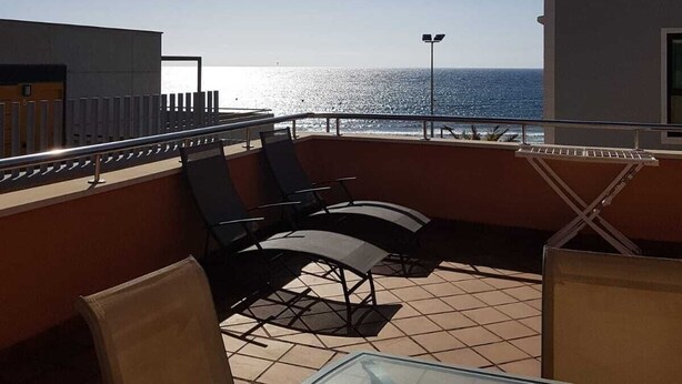 A sunlit outdoor seating area with a glass table and chairs, overlooking a calm sea.