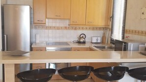 A kitchen with wooden cabinets, a stainless steel refrigerator, a kettle, and a toaster. There are three bar stools with metal bases.