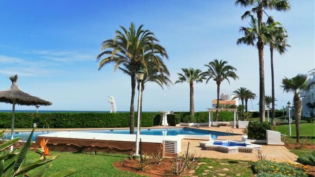 Ein Poolbereich mit Palmen, einem Reetdachschirm und Blick auf das Meer.