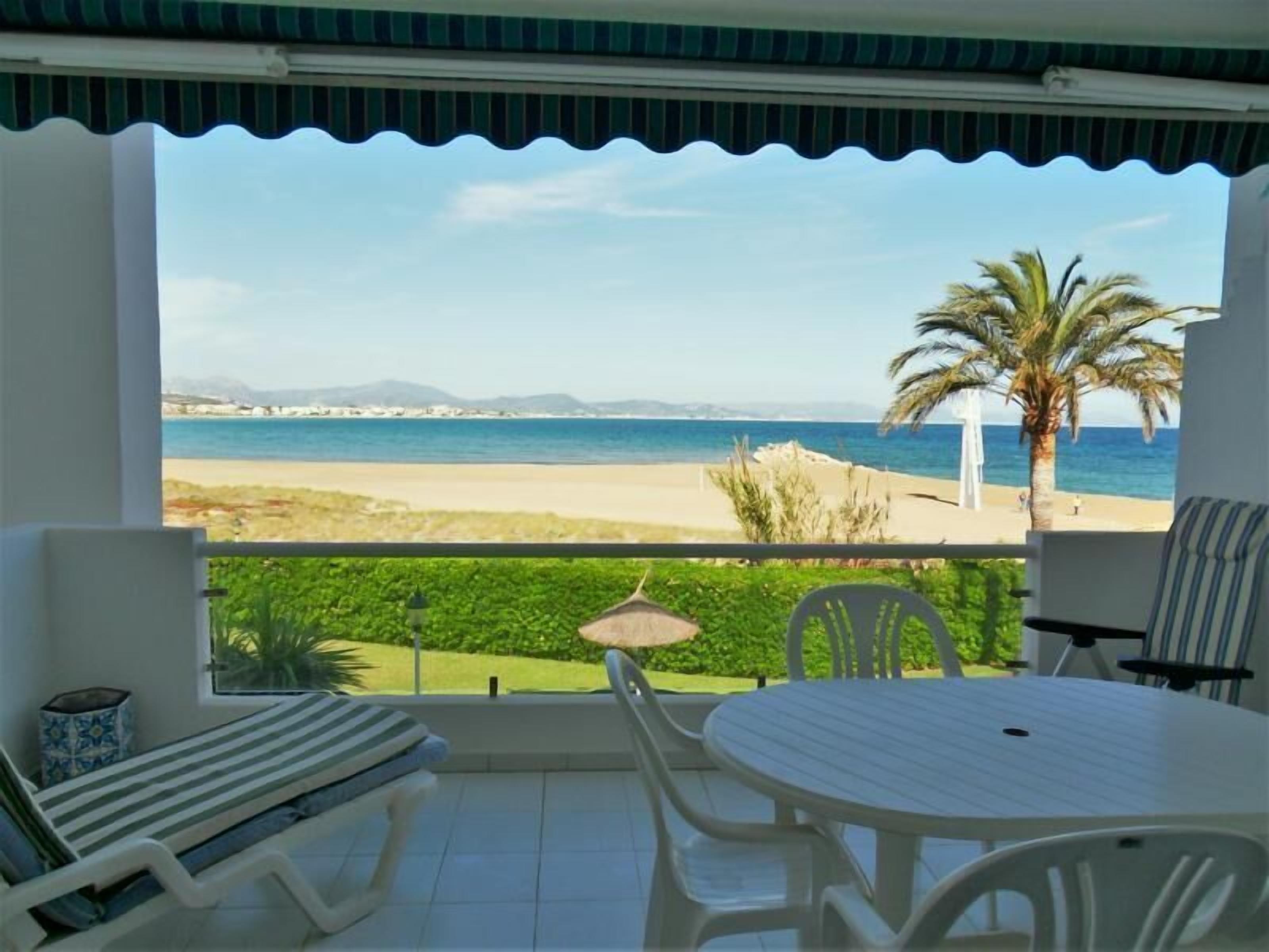 A balcony with a view of the beach, a table, chairs, and a cushioned bench.