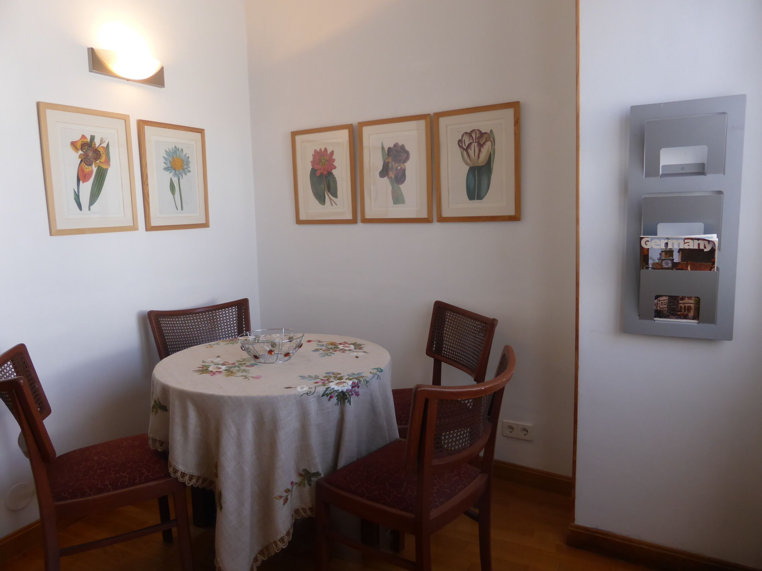 A dining area with a round table covered by a floral tablecloth, surrounded by four wooden chairs. There are framed floral artworks on the wall above the table.
