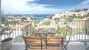A balcony with a wooden table and chairs, overlooking a coastal town with buildings and a sea view.