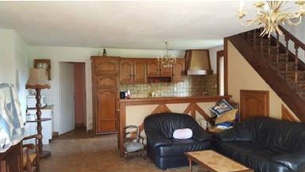 A living room with a black leather sofa, a wooden coffee table, and a kitchen area with wooden cabinets and a chandelier. - Very quiet family holiday home (Isigny-le-Buat)