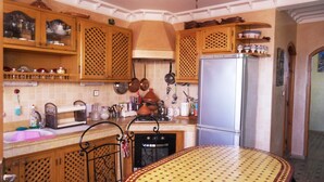 Une cuisine avec des meubles de rangement en bois, un réfrigérateur et une table à manger recouverte d’une nappe à motifs.