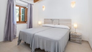 Un llit doble amb un capçal de fusta, dues taules de nit amb làmpades, una finestra amb cortines i un sòl de rajoles.