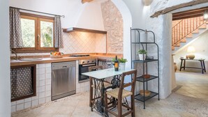 Una cuina amb paret de pedra, bigues de fusta, salabost d'azulejos, un rentapeus, una cuina i una zona de menjador amb taula i cadires.